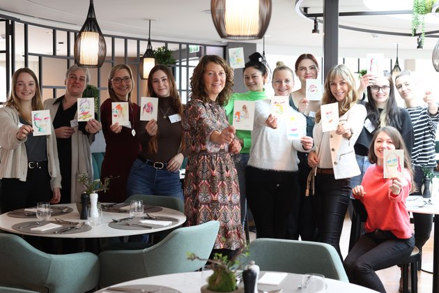 Group of 13 women in a modern restaurant holding colorful drawn cards to the camera and smiling.