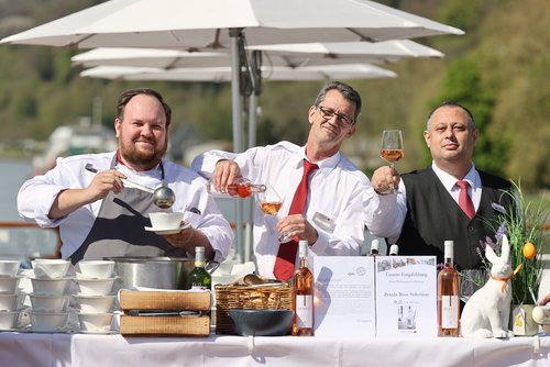 Three men in work clothes at a table with soup bowls, wine bottles, and a white rabbit decoration, one ladling soup, another pouring rosé wine into a glass.