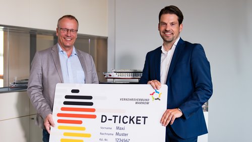 Jörg Eichler and another man are holding a large model of a D-ticket from the Warnow Transport Association with the name Maxi Muster and customer number 1234567.