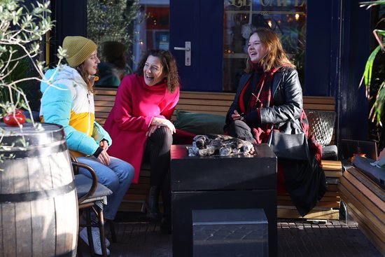 Three women are sitting outside on a wooden bench and laughing, one is wearing a colorful jacket and a yellow hat, another a pink coat, and the third a black leather jacket and a red scarf.