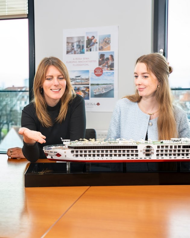 Two women are sitting at a table and looking at a detailed model of a river cruise ship in a bright room with windows and posters in the background.
