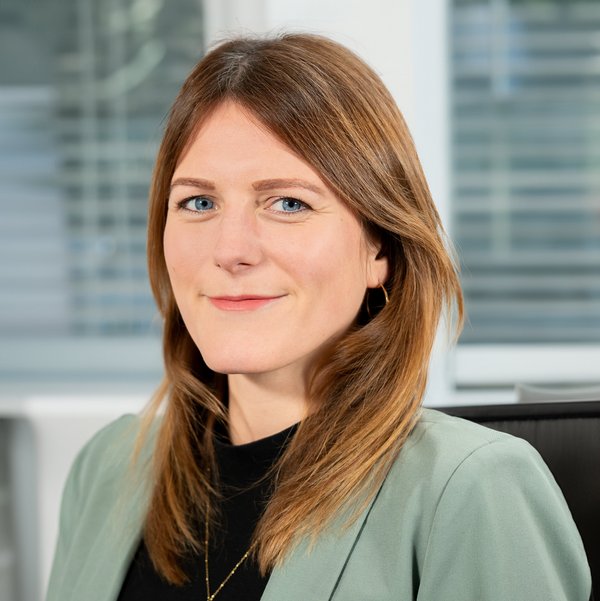 Nadine Richter, with shoulder-length brown hair, wearing a green blazer and black blouse, smiles slightly in front of office windows.