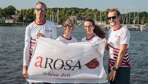 Four A-ROSA employees, including Stefan Sprunk, are holding a white flag with the inscription A-ROSA Schöne Zeit in front of a lake with boats in the background.