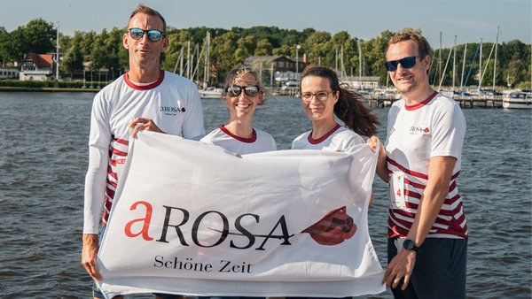 Four A-ROSA employees, including Stefan Sprunk, are holding a white flag with the inscription A-ROSA Schöne Zeit in front of a lake with boats in the background.