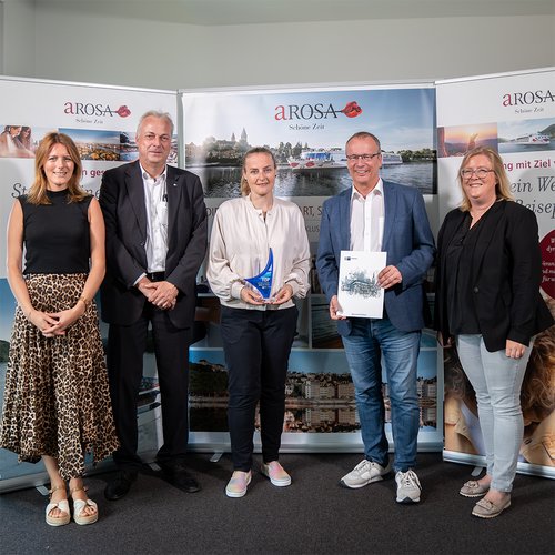 Five people, including Jörg Eichler, stand in front of banners with the logo A-ROSA Schöne Zeit. Two are holding an award and a certificate for top training company.