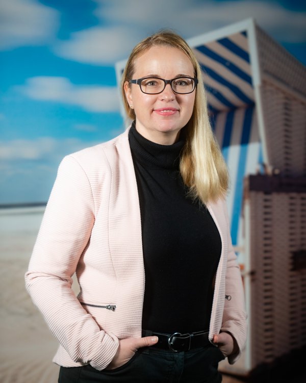 Anne Riesner-Koch with glasses, blonde hair, black turtleneck sweater, and light blazer in front of a beach chair and blue sky.