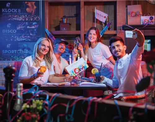 Four young adults are sitting at a table, clinking glasses and taking a selfie, with billboards in the background displaying the words Klock 8, Disco, and Sports Club. One of them is holding a large LOKA card in their hand.