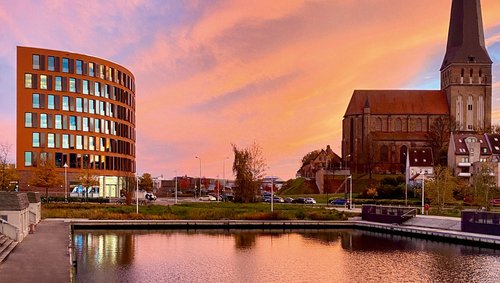 View of the A-ROSA office building in Rostock next to a church with a tall tower at sunset, water in the foreground reflecting the orange sky.