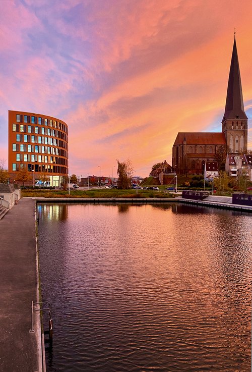 View of the A-ROSA office building in Rostock next to a church with a tall tower at sunset, water in the foreground reflecting the orange sky.