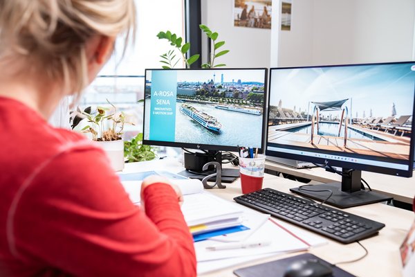 A woman in a red sweater is working at a desk with two monitors, one of which shows a river cruise ship with the text A-ROSA SENA Innovatives E-Motion Ship.