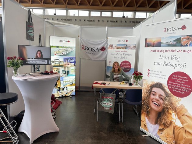 A-ROSA booth with banners about careers and training in the travel sector, woman sitting at the table, monitor showing video with subtitles, roses on tables