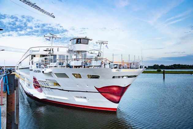 The river cruise ship A-ROSA SENA with a white hull and red lip motif on the bow is docked in the harbor with calm water and a cloudy sky.