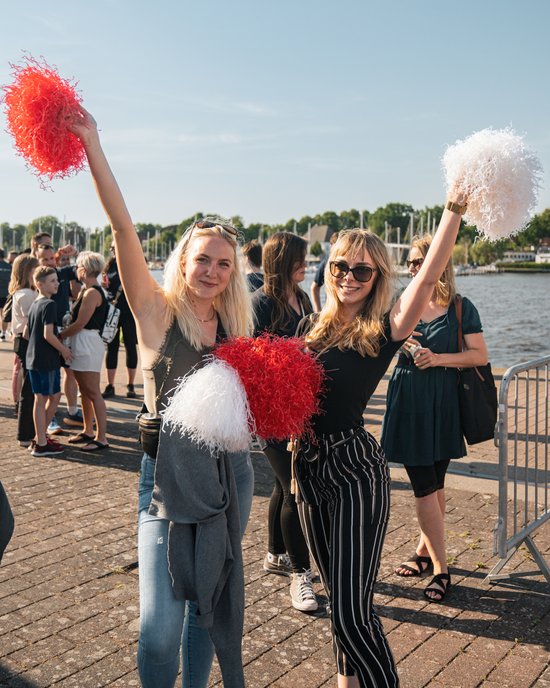Two young women with red and white pom-poms are celebrating by the water in sunny weather, with more people and boats in the background.