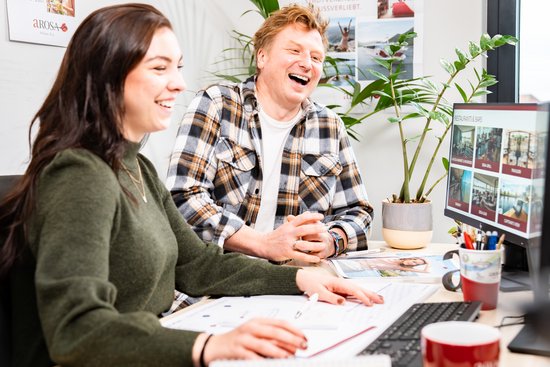 Two laughing people are sitting at a desk with a computer and documents, with plants and posters in the background.