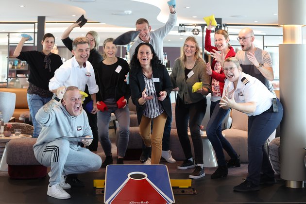 Eleven people are playing cornhole indoors, throwing colorful bags onto a game board and showing joy and enthusiasm.