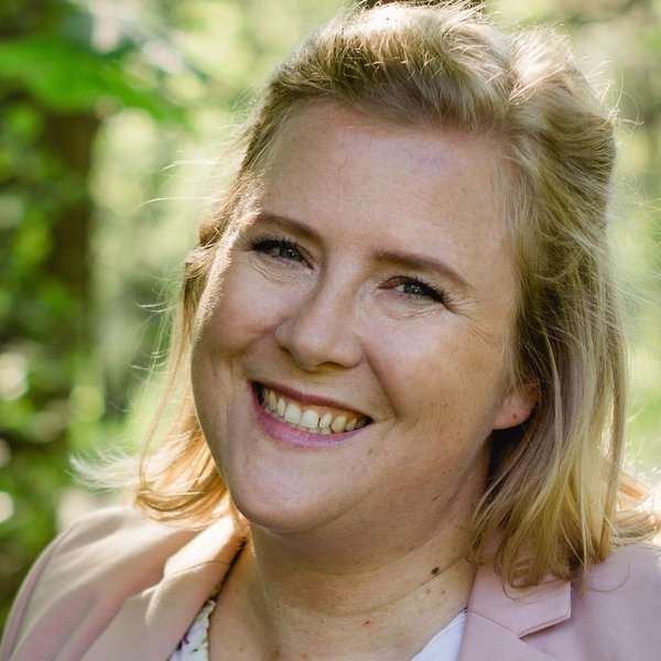 Smiling woman with blonde hair, wearing a light pink blazer, in front of a green, blurred background.
