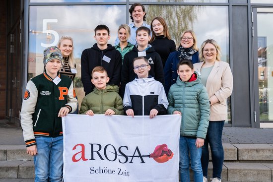 Group photo of young people and adults in front of a building, with four boys at the front holding a banner with the inscription A-ROSA Schöne Zeit.