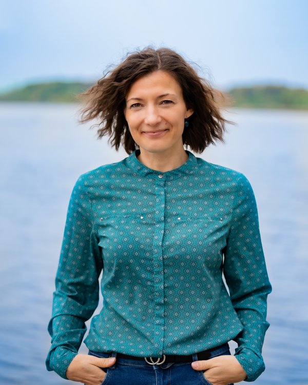 A woman with shoulder-length brown hair blowing in the wind is wearing a turquoise patterned shirt and standing in front of a body of water with a blurred background.