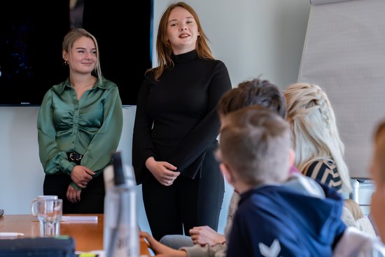 Two young women stand in front of a group of children in a classroom, one wearing a black top, the other a green shirt.