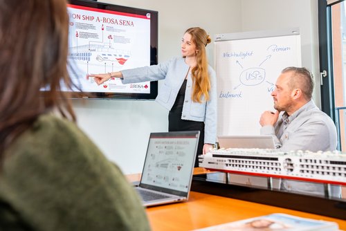 A woman presents a cruise ship infographic on a screen, two people are listening, a model ship is on the table.