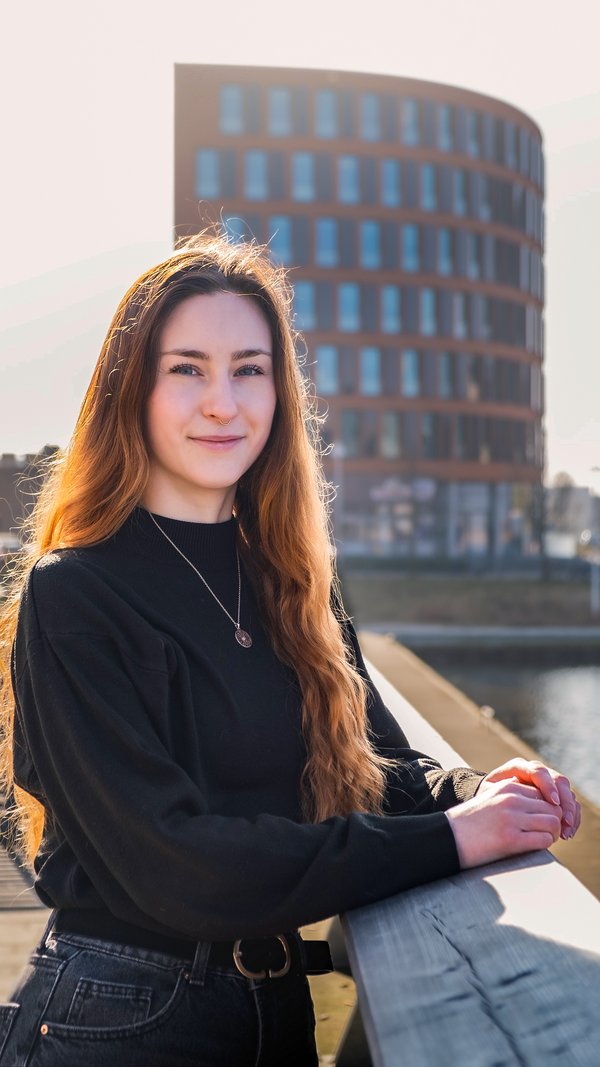 Young woman with long red hair and a black sweater leans against a railing in front of a modern, round office building by the water.