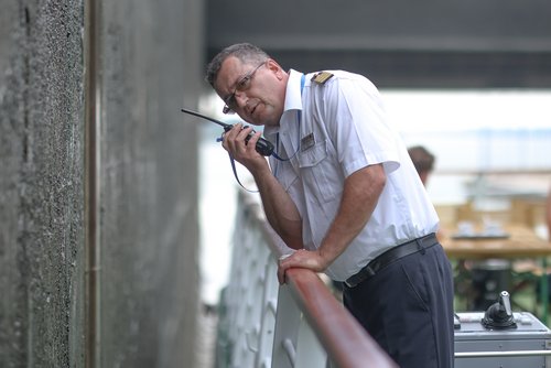 Man in a white shirt with epaulettes speaks into a walkie-talkie and leans against a railing