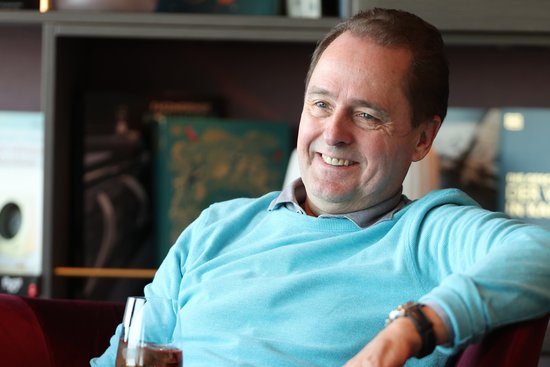 Man in a light blue sweater smiles relaxedly sitting in front of a bookshelf.