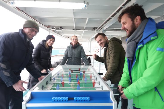 Five people in winter clothing are playing table football on a covered outdoor deck.