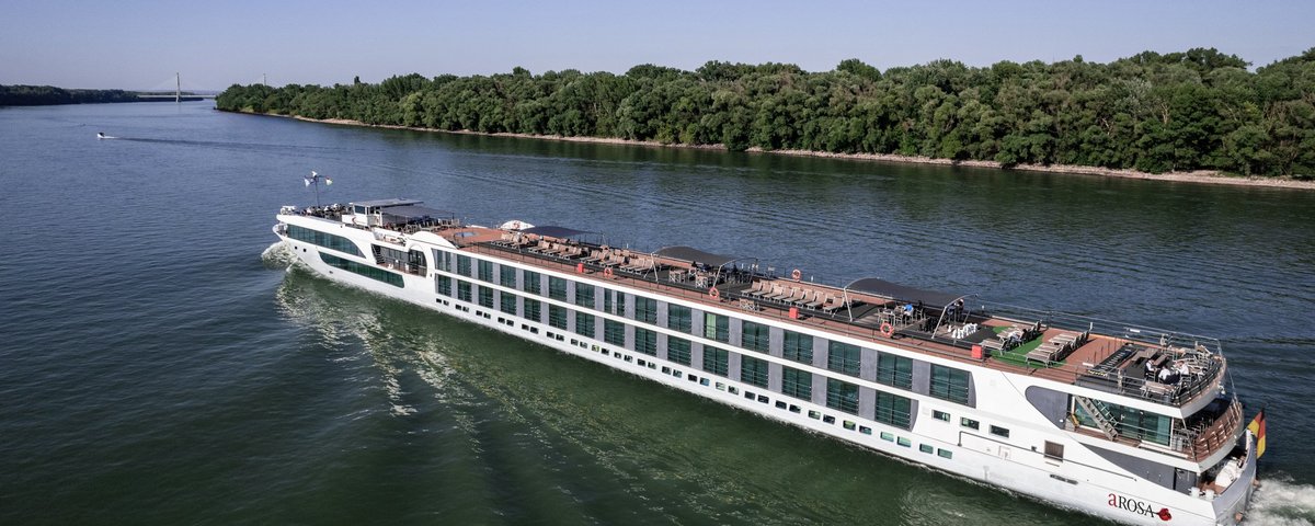 A river cruise ship is sailing along a wooded bank on the Rhine. The sun deck is equipped with a whirlpool, sun loungers, and parasols.