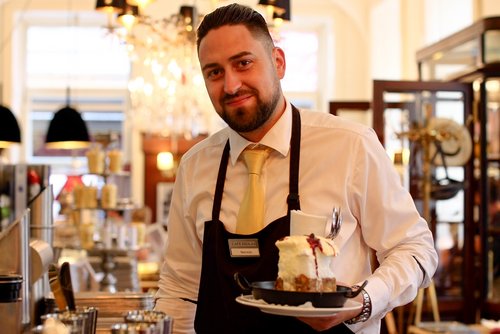Waiter with a black apron and yellow tie holding a plate with a piece of cream cake in a café