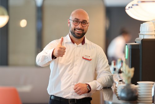 Man in a white shirt with a name tag gives a thumbs up next to a coffee machine and white dishes.