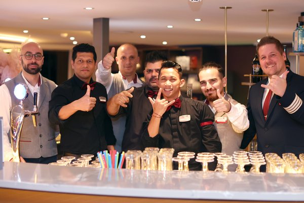 Seven male bartenders and waiters in black and gray work attire with red bow ties and ties stand behind a bar with glasses and colorful straws.