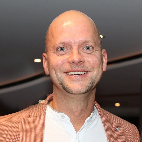 Bald man smiling, wearing a white shirt and a red jacket, in front of a blurred background.