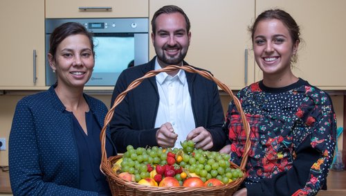 Three smiling people in a kitchen are holding a large basket with grapes, strawberries, and other fruits together.