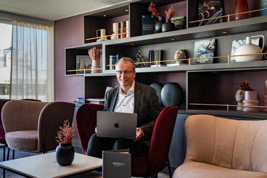 Jörg Eichler sits in a modern office on a red chair with an Apple laptop on his lap, surrounded by comfortable chairs and decorative shelves with books and vases.