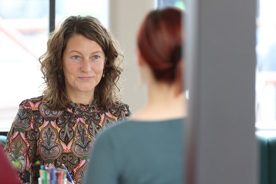 A woman with curly brown hair and a patterned top sits across from another woman with red hair in a bright room.