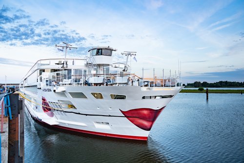 The river cruise ship A-ROSA SENA with a white hull and red lip motif on the bow is docked in the harbor with calm water and a cloudy sky.
