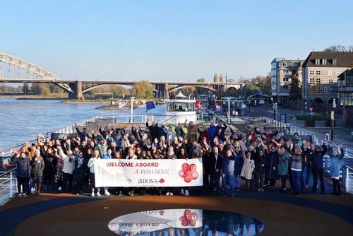 Large group of people on a river cruise ship holding a banner that reads WELCOME ABOARD A-ROSA SENA! and waving, with a bridge and riverbank in the background.