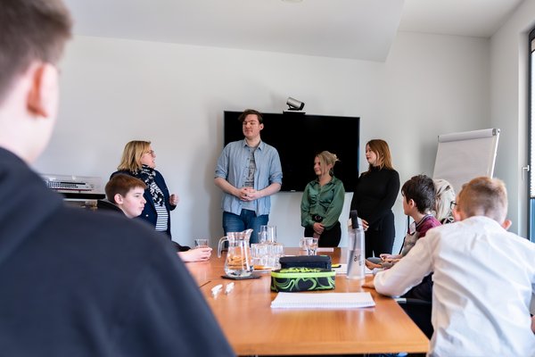 Teenagers are sitting at a conference table, three people are standing in front of a screen and speaking in a bright meeting room.