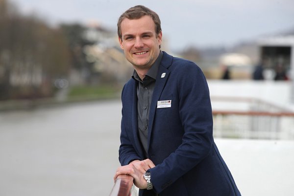Alexander Steube leans smiling against a railing, wearing a dark blue blazer and shirt, with a blurred river and city in the background.
