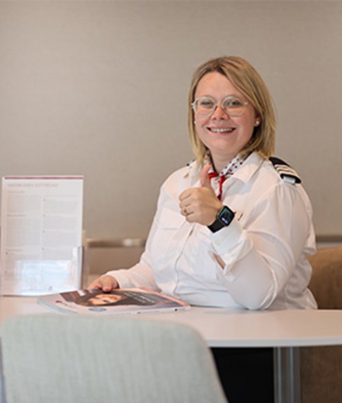 Smiling woman in a white uniform with shoulder insignia and glasses sits at a table and gives a thumbs up.